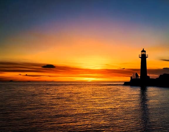 夕暮れか夜明け前の、穏やかな水面に佇む灯台から、柔らかく温かい光がまっすぐに放たれている風景