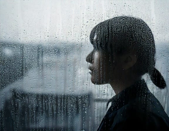 雨の日に、窓ガラスについた水滴の向こう側を、ぼんやりと眺めている人物のシルエット